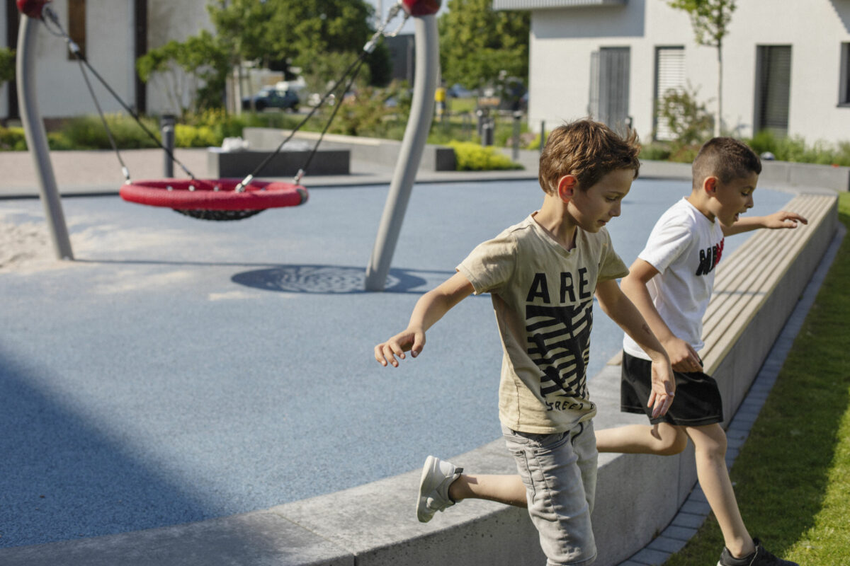 Zwei Kinder spingen zusammen auf einem Spielplatz von einer kleinen Mauer auf den Rasen.