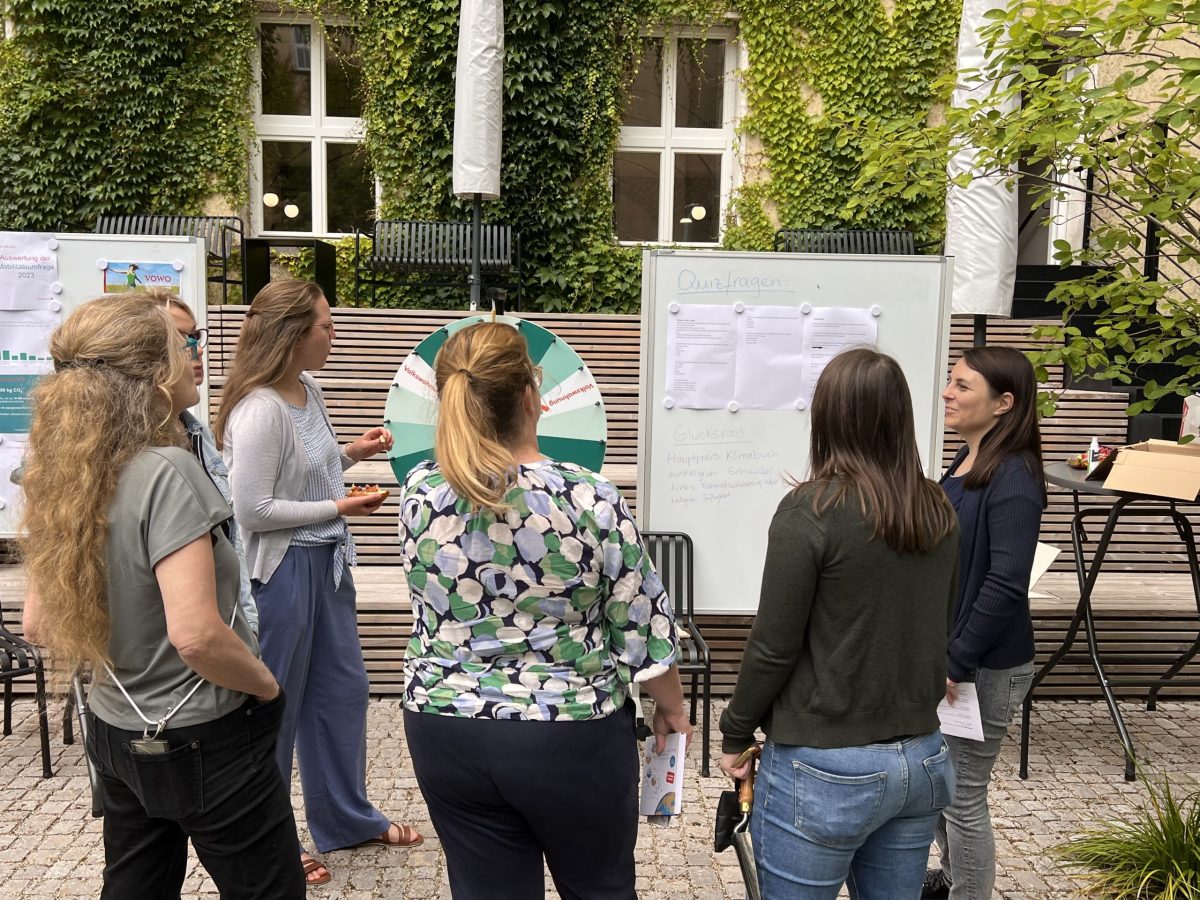 Personen stehen um ein Glücksrad und eine Flipchart mit Informationen und tauschen sich aus.