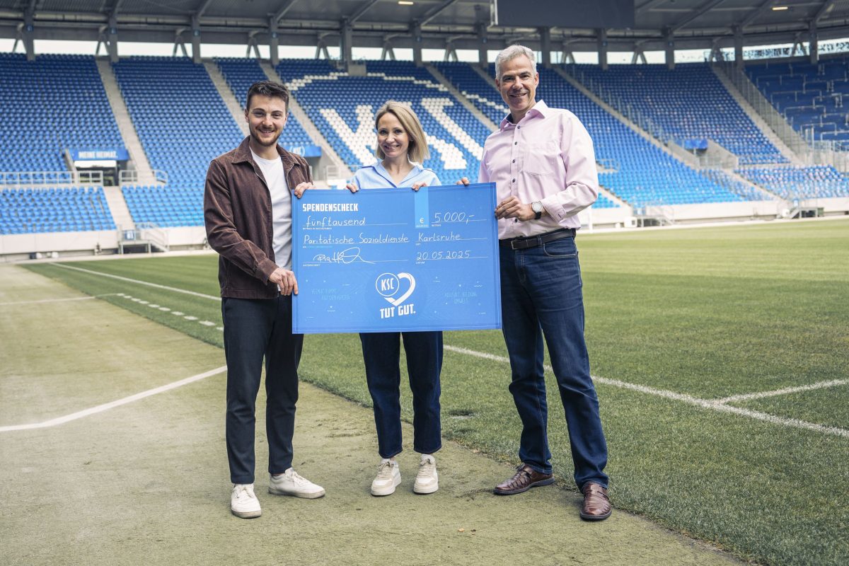Drei Personen stehen im Fußballstadion des KSC auf dem Spielfeld und halten einen Spendenscheck über 5.000 Euro für die Paritätischen Sozialdienste.