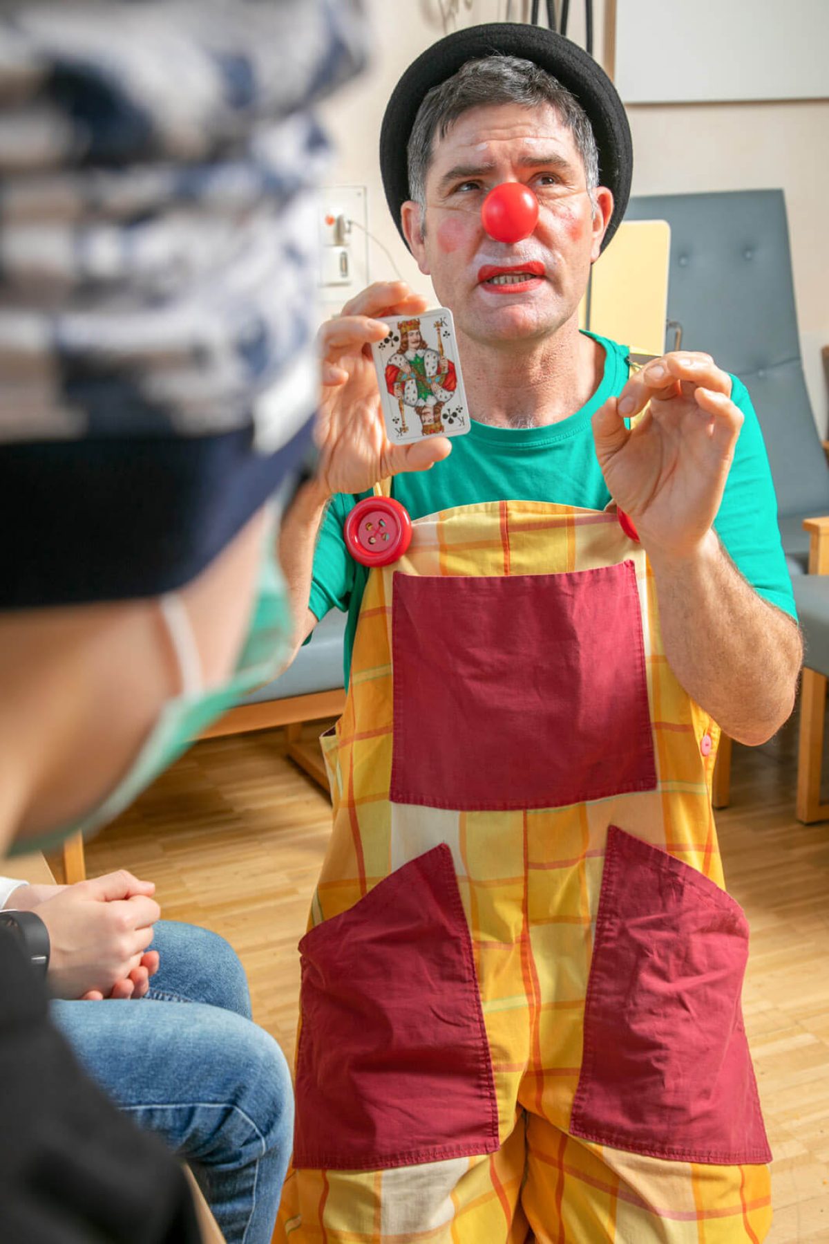 Ein Klinikclown mit roter Nase, Hut und kariertem Kostüm zeigt einem Kind in einem Behandlungsraum eine Spielkarte.