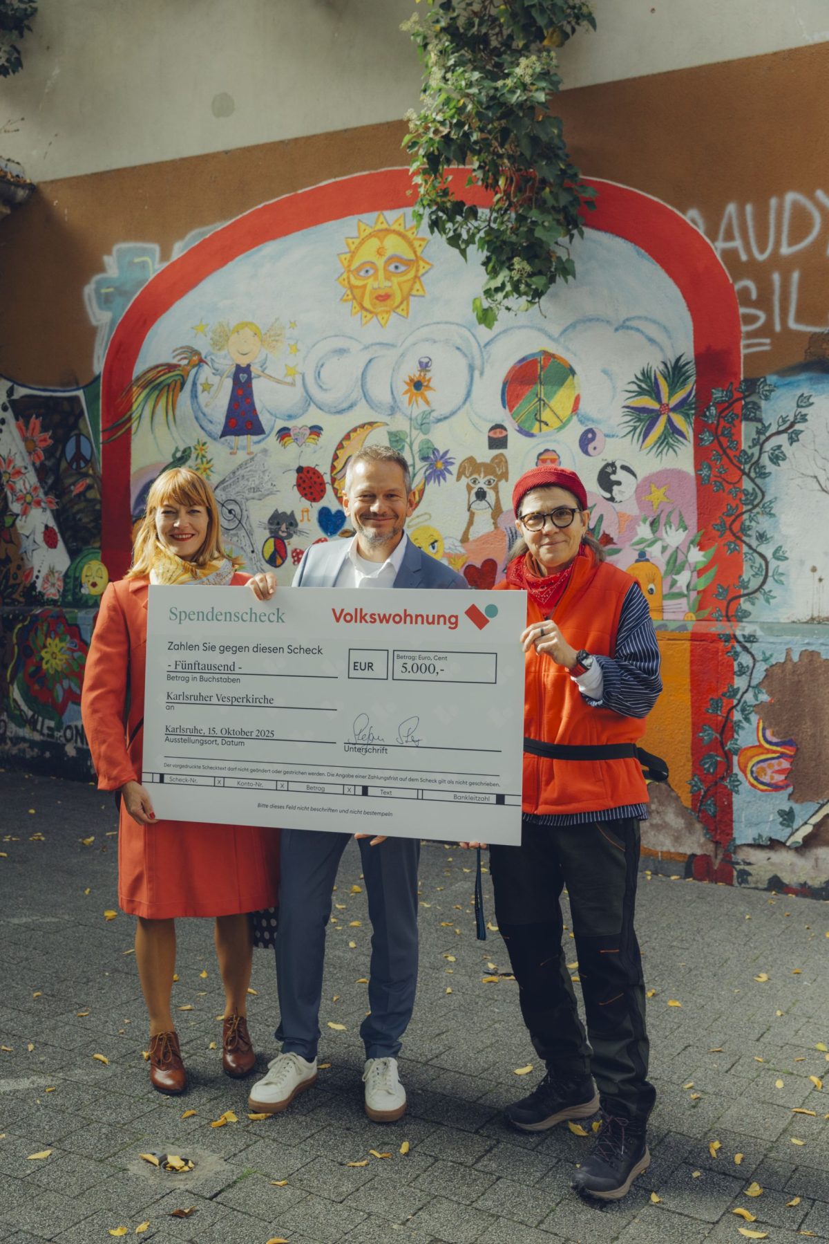 Drei Personen stehen vor einer bunt bemalten Wand und halten einen Spendenscheck der Volkswohnung über 5.000 Euro für die Karlsruher Vesperkirche.