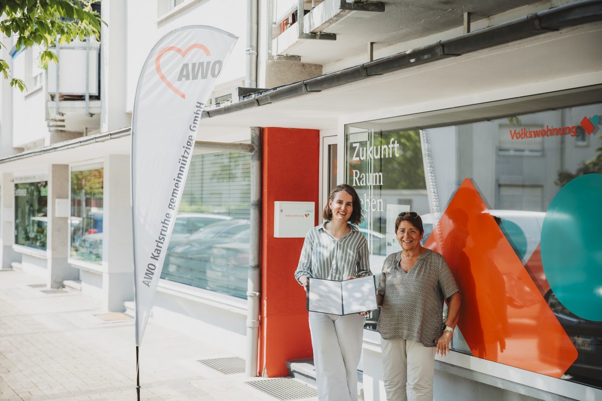 Zwei Frauen stehen vor einem Volkswohnung-Standort in Mühlburg neben einer AWO-Fahne; eine hält eine geöffnete Vertragsmappe in den Händen.